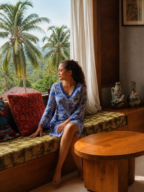 A person sitting on a cushioned bench by a window with a view of palm trees. - Villa OMantra Home Bali (Sukasada)