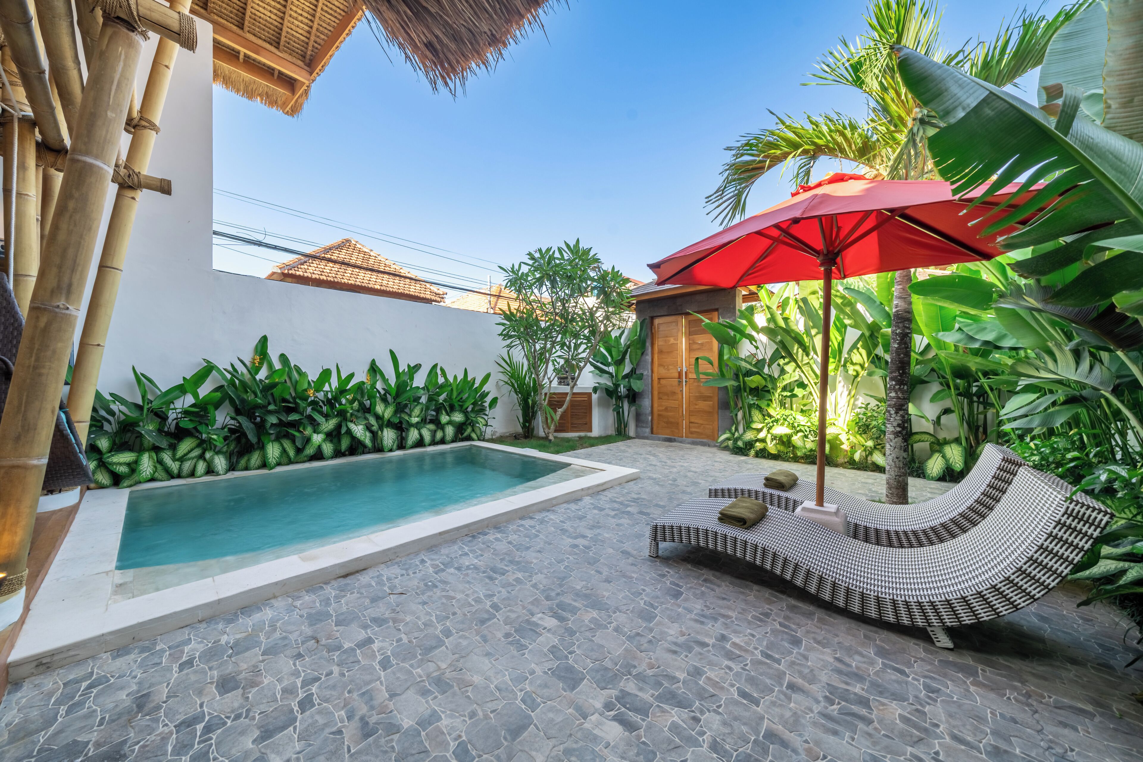 A swimming pool with a stone floor, surrounded by lush greenery and a red umbrella.
