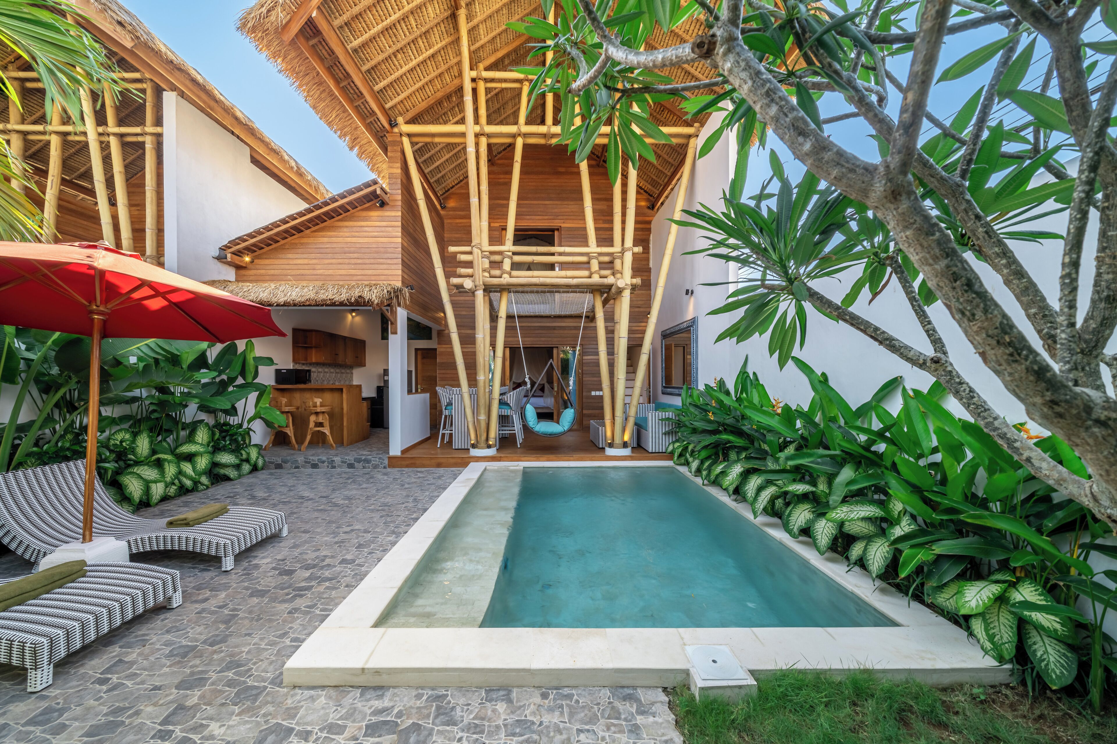 Un área de piscina con sillones, una sombrilla roja y una estructura de techo de paja.
