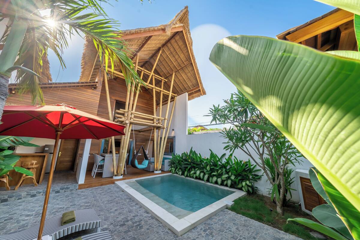 A pool area with a thatched-roof structure, outdoor seating, and lush greenery.
