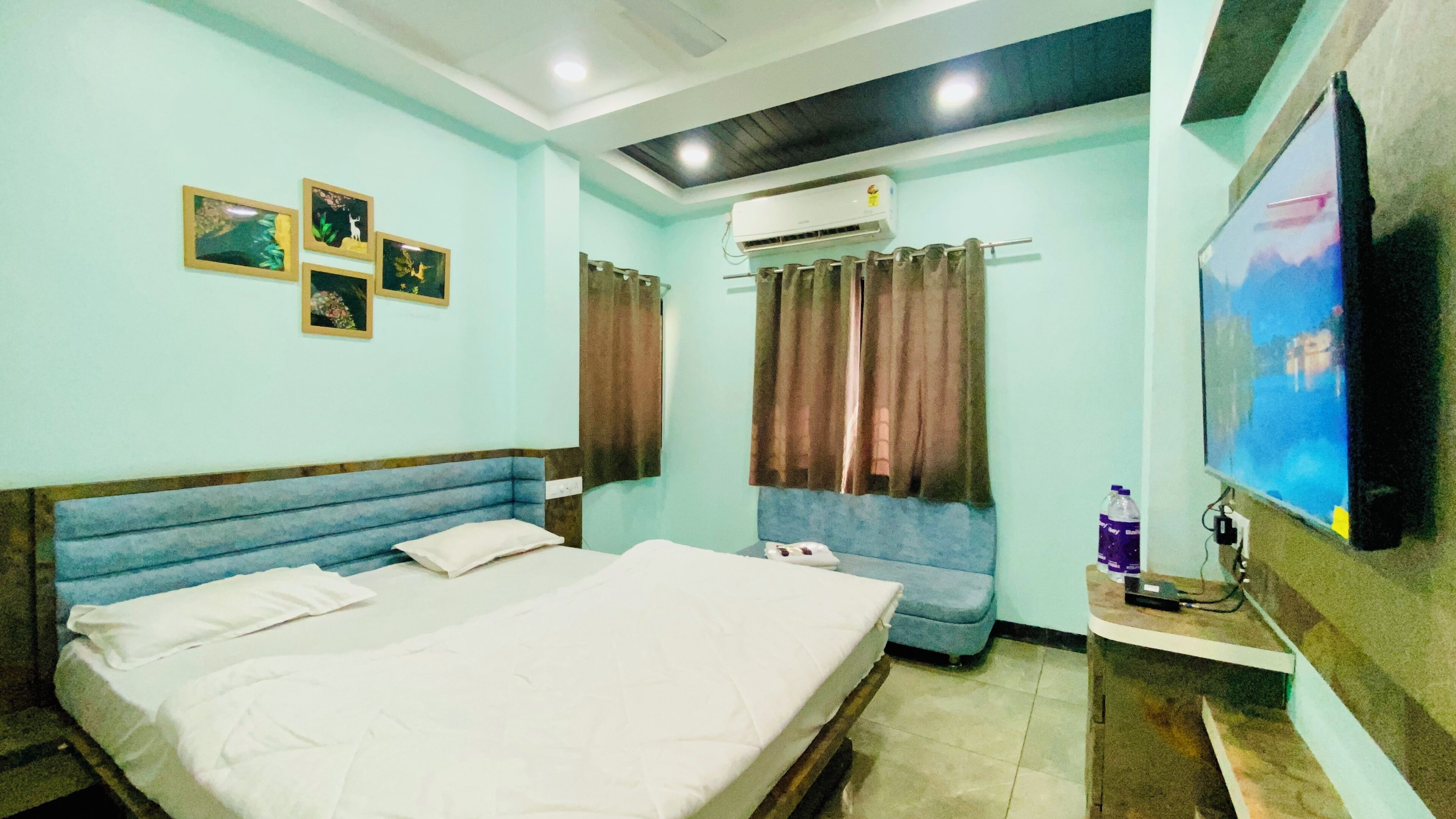 A hotel room with a bed, a small sofa, a TV, an air conditioner, and a wall-mounted TV.