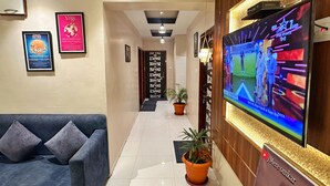 Rebuda d'hotel amb taulell de recepció, un gran televisor de pantalla plana que mostra un esdeveniment esportiu, pòsters emmarcats a les parets, un sofà blau i una planta en test.