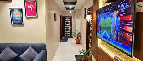 A hotel lobby with a reception desk, a large flat-screen TV displaying a sports event, framed posters on the walls, a blue sofa, and a potted plant.