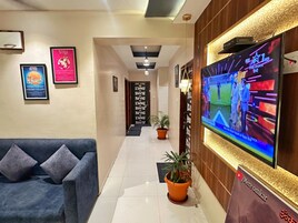 Um saguão de hotel com balcão de recepção, uma grande televisão de tela plana exibindo um evento esportivo, pôsteres emoldurados nas paredes, um sofá azul e um vaso de plantas.