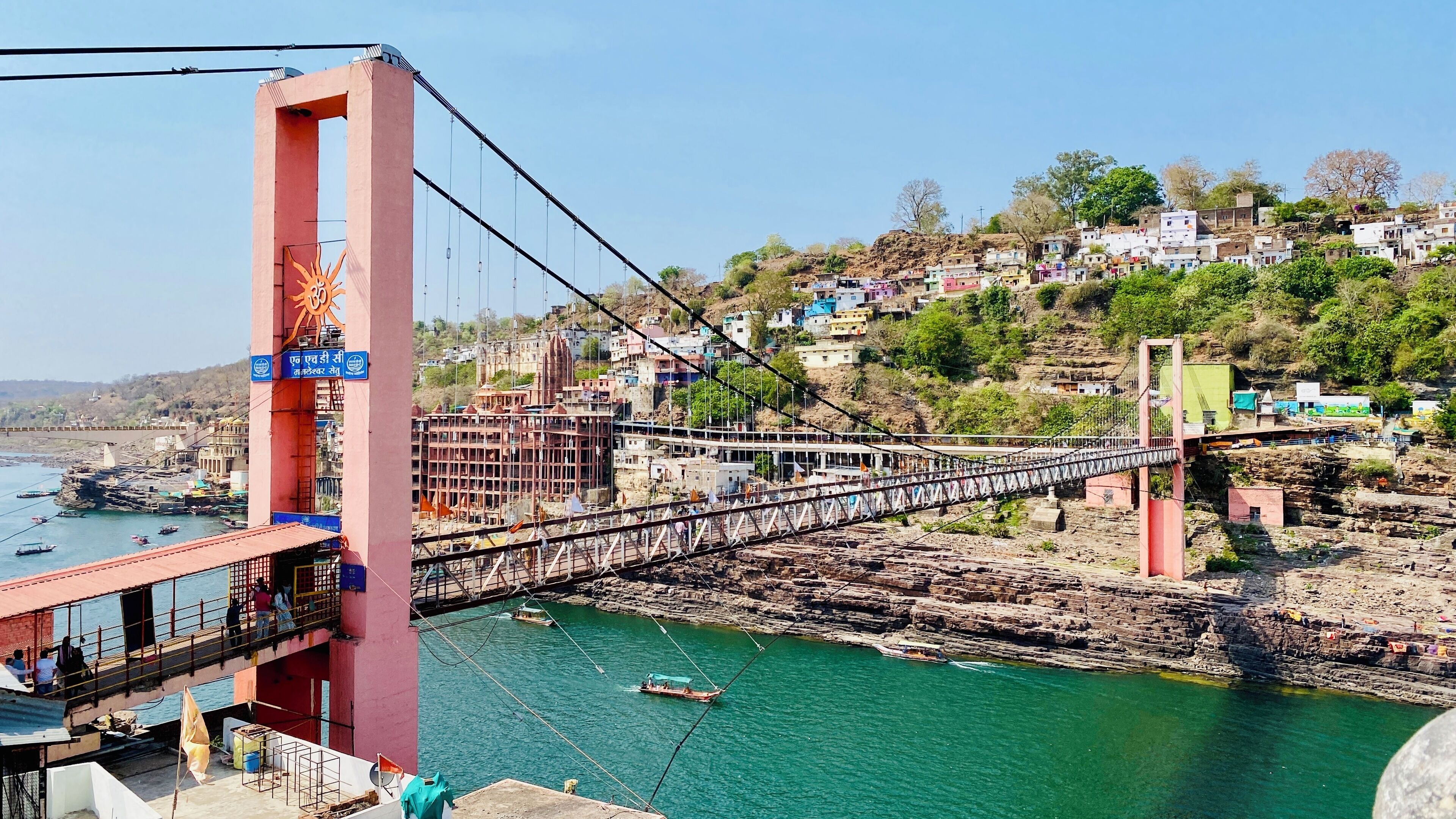 A bridge with a large tower and a river below. There are boats on the river and buildings on the opposite bank.