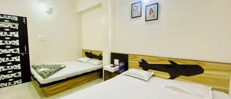 A hotel room with two single beds, a wooden headboard, a small bedside table with a remote control, and two framed pictures on the wall.