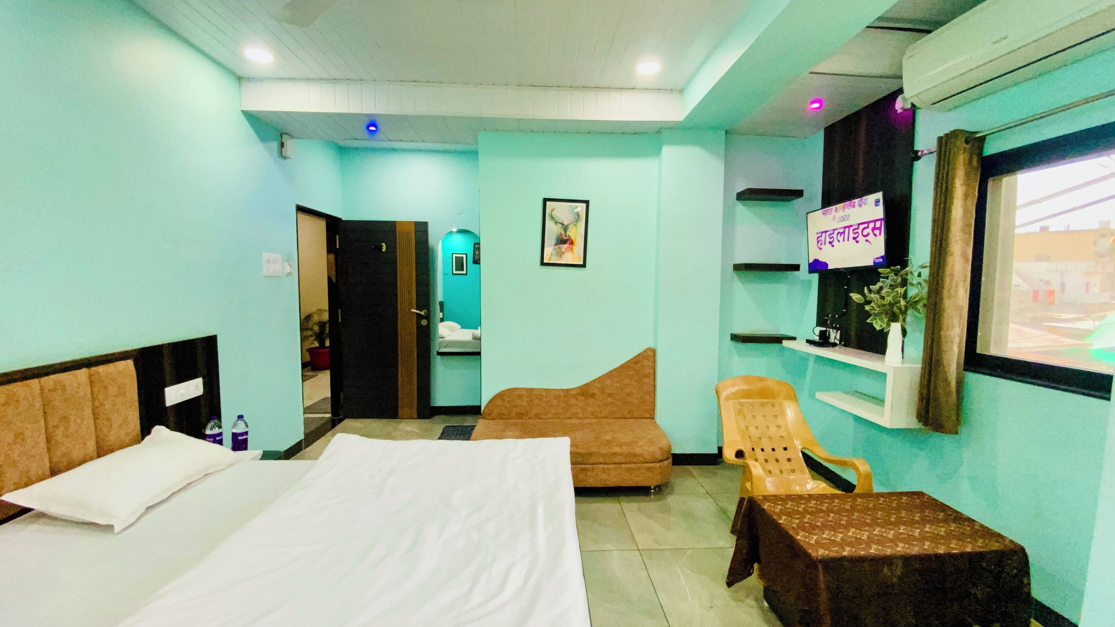 A hotel room with a bed, a sofa, a small table, a chair, a mirror, a shelf with a plant, and a window.