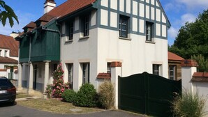 Une maison à deux étages, avec une façade blanche et des éléments décoratifs sombres, un toit recouvert de tuiles rouges et un portail vert.