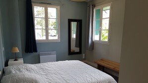 Une chambre à coucher avec un lit, une table de chevet avec une lampe, un banc, un radiateur et une fenêtre avec des rideaux.