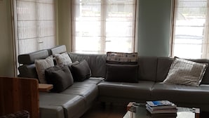Un salon comprenant un canapé d’angle, des fauteuils en osier, une table basse en verre et de grandes fenêtres avec des rideaux.