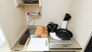 Uma kitchenette compacta com pia, tábua de corte, frigideira, balança e uma pequena prateleira com uma xícara e um recipiente.