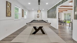 Um espaço interior moderno com mesa de bilhar, uma grande letra decorativa 'X', e uma área de refeições visível por uma porta aberta.