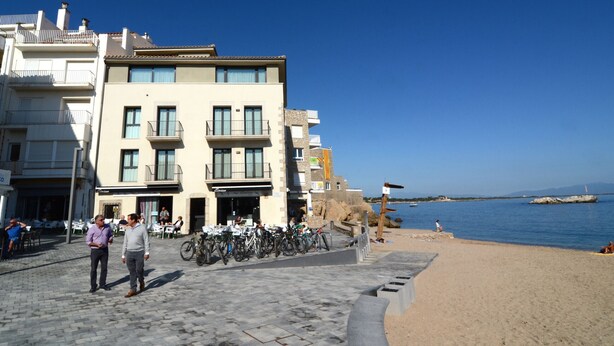 Un bâtiment situé sur la côte, doté de balcons, d’une promenade dallée et avec une plage en arrière-plan.