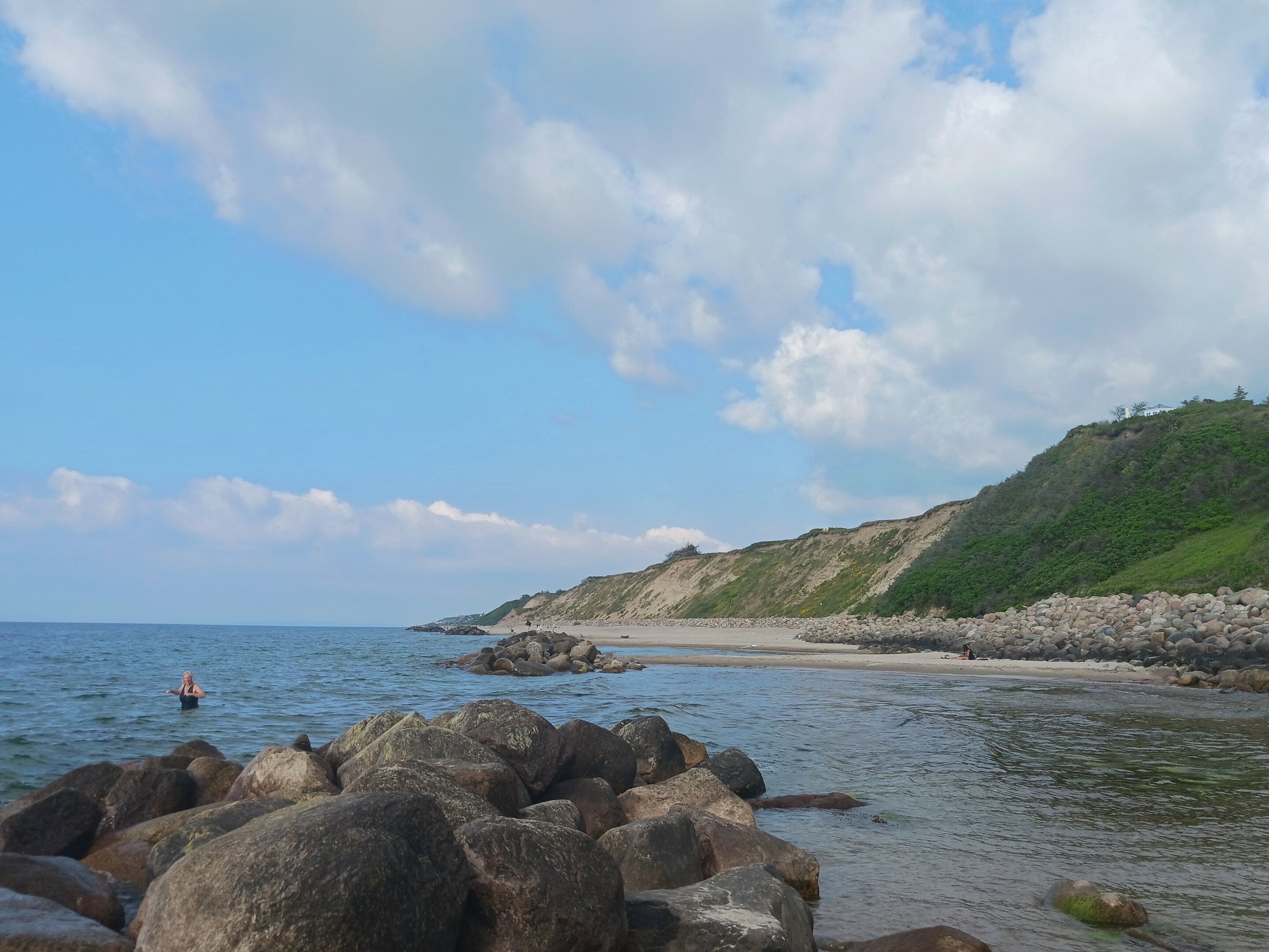 Paisagem costeira com grandes rochas em primeiro plano, uma praia arenosa e uma colina com vegetação.