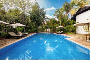 A rectangular swimming pool with blue tiles, surrounded by lounge chairs and umbrellas, set against a backdrop of lush greenery and a clear sky. - Angkor Green Boutique (Siem Reap)