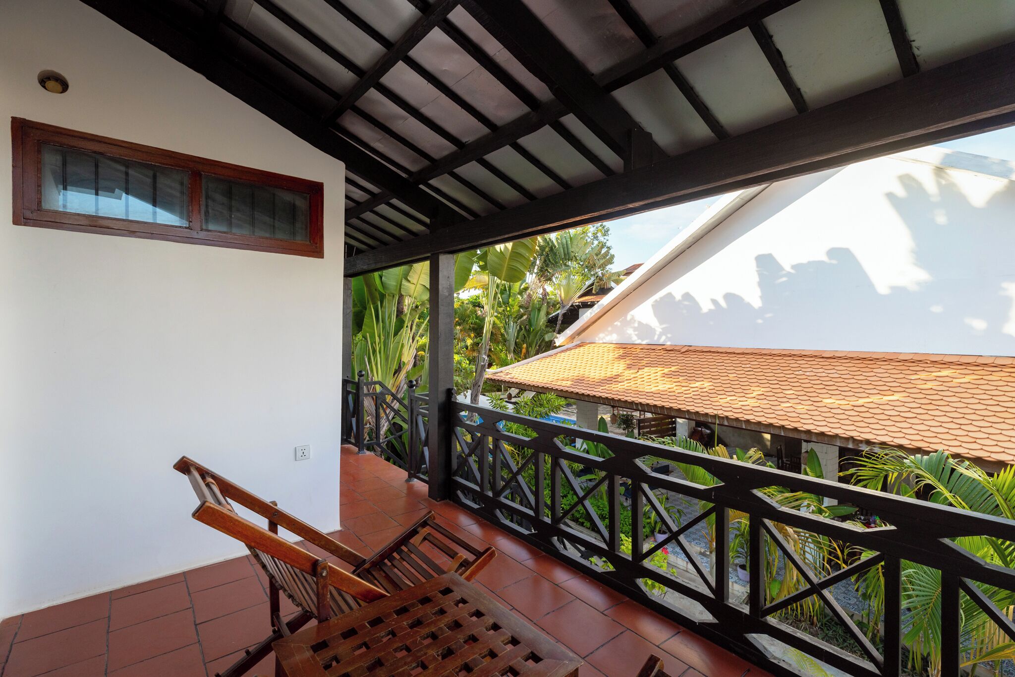 Varanda com cadeiras de madeira, chão de azulejos, janela e vista para um jardim com uma construção de telhado.