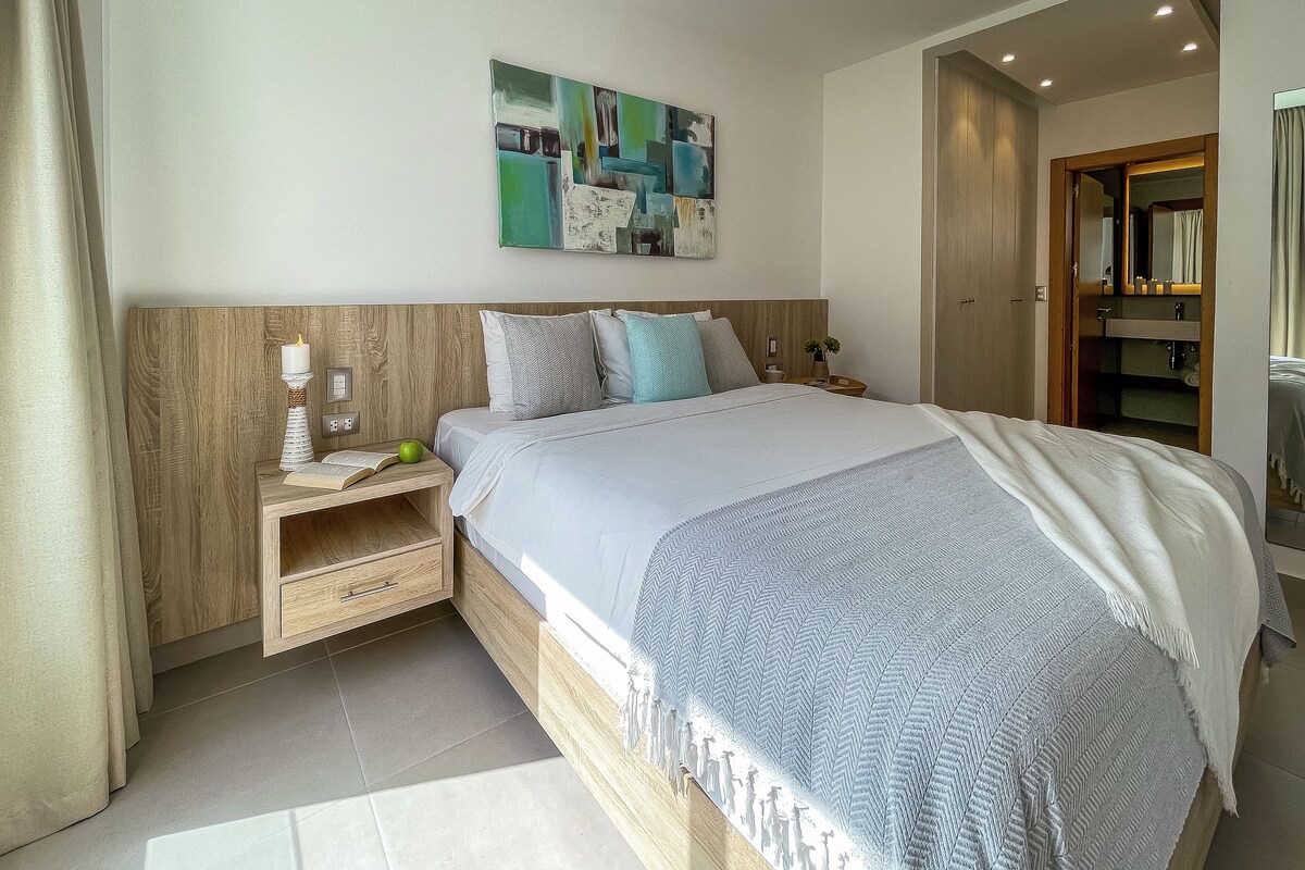 A modern bedroom with a wooden headboard, a large bed with a grey and white patterned blanket, a bedside table with a book and a green apple, and a wall art piece above the bed.