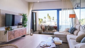 A modern living room with a flat-screen TV, a sofa, a dining area, and a balcony with a view of palm trees. - Beach Homes Punta Cana (Punta Cana)