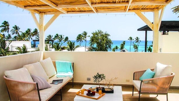 A balcony with wicker furniture, a coffee table with fruits and candles, and a view of the ocean and palm trees. - Beach Homes Punta Cana (Punta Cana)
