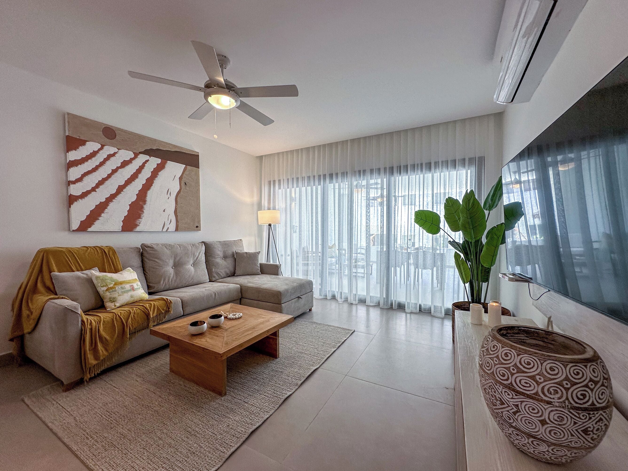 A modern living room with a sectional sofa, a wooden coffee table, a large plant, and a ceiling fan.