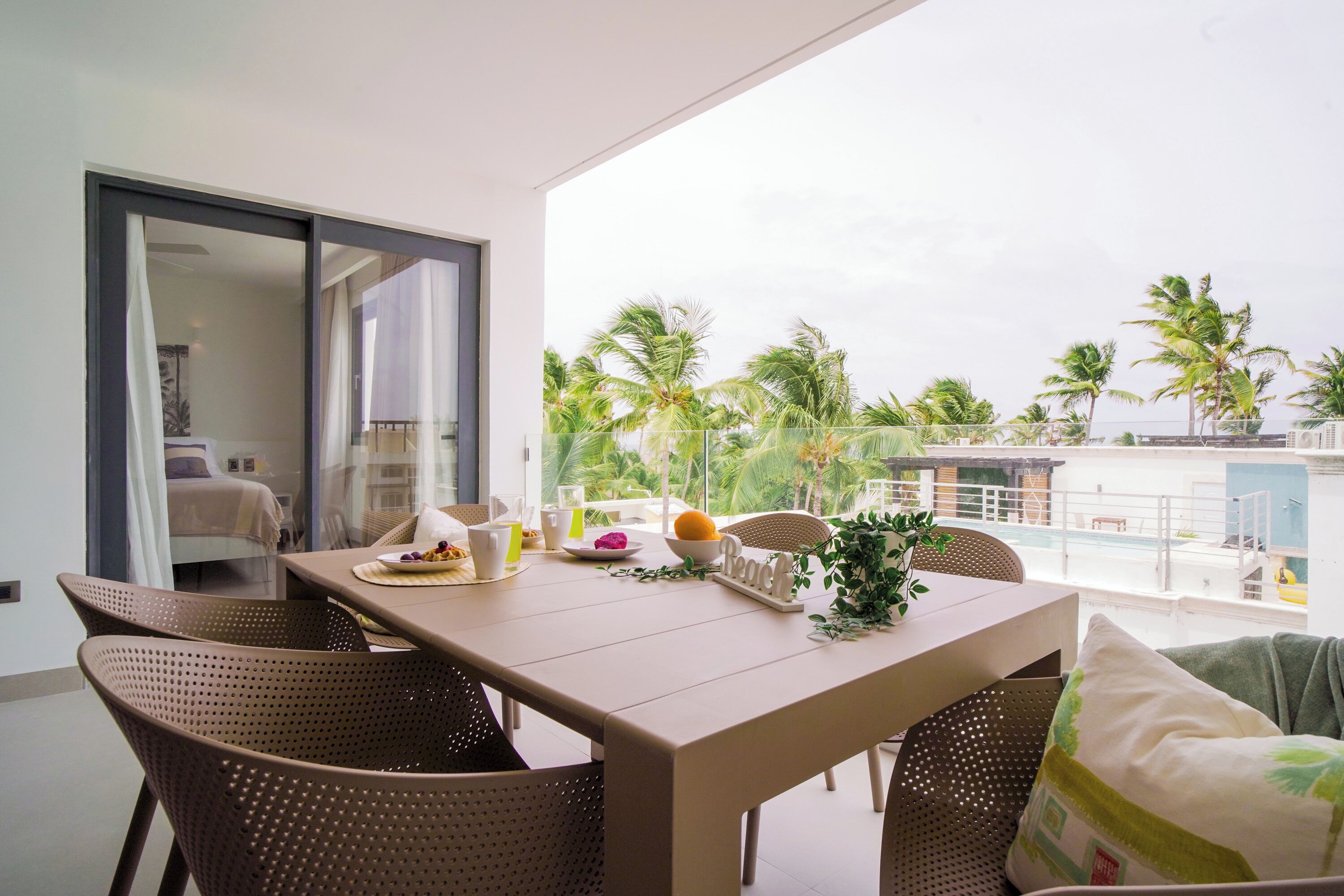 Un balcón con mesa y sillas de mimbre para dos personas, con vista a un paisaje tropical con palmeras y una piscina.