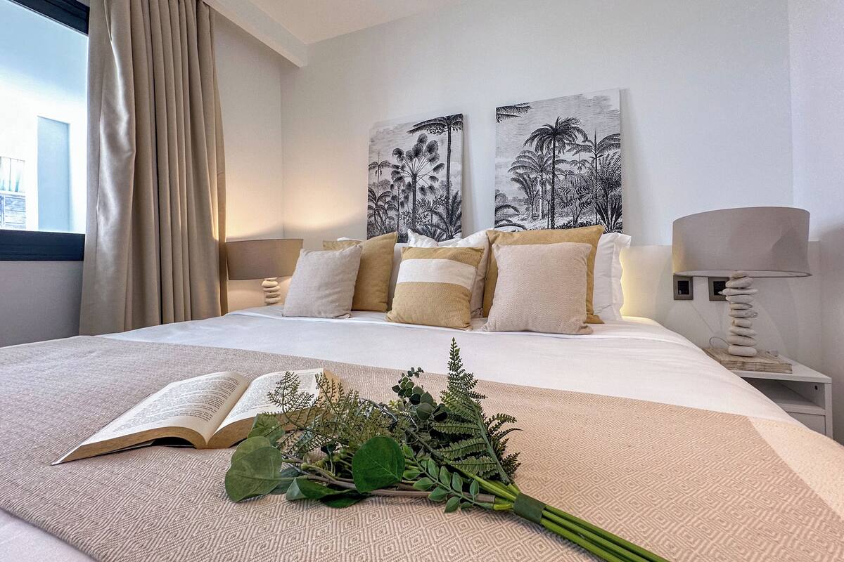Una cama bien hecha con un libro, un ramo de flores, una lámpara y dos cuadros con palmeras.
