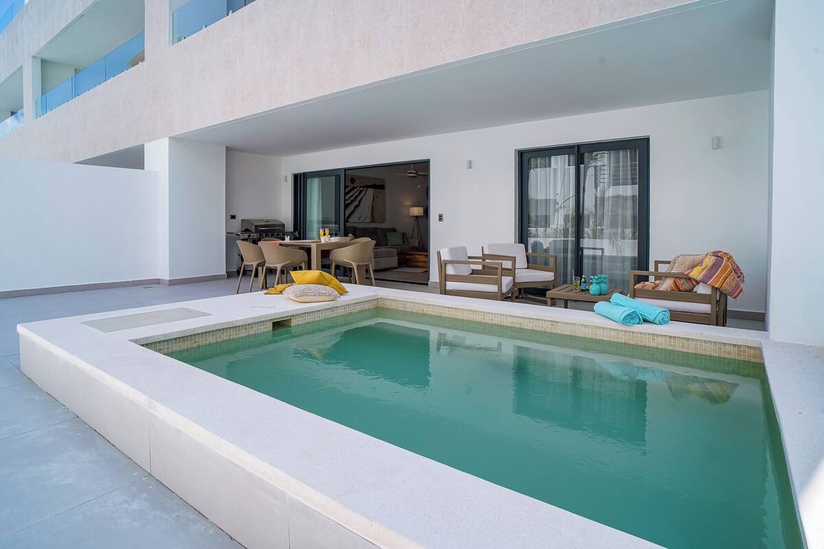 Un área de piscina exterior moderna con una piscina rectangular, sillas de descanso y una mesa de comedor.