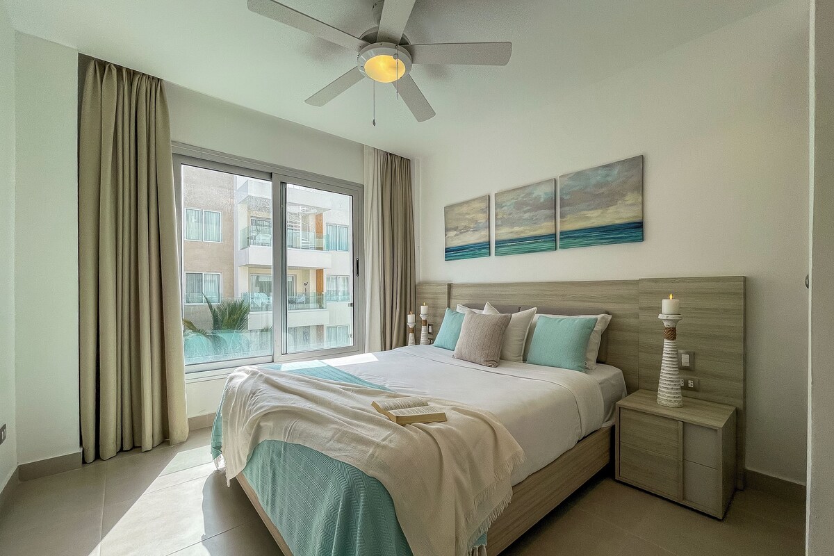 A bedroom with a large bed, a ceiling fan, a nightstand with a candle, and a view of a building through the window.