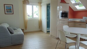 A modern living room with a grey sofa, a dining table with white chairs, a kitchen with white cabinets, and a skylight.