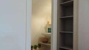 A narrow hallway with a white wall, a wooden floor, and a small shelf with a basket and a roll of toilet paper.