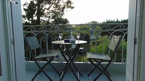 A balcony with a table and two chairs, a car parked in the background, and a view of trees and a building.