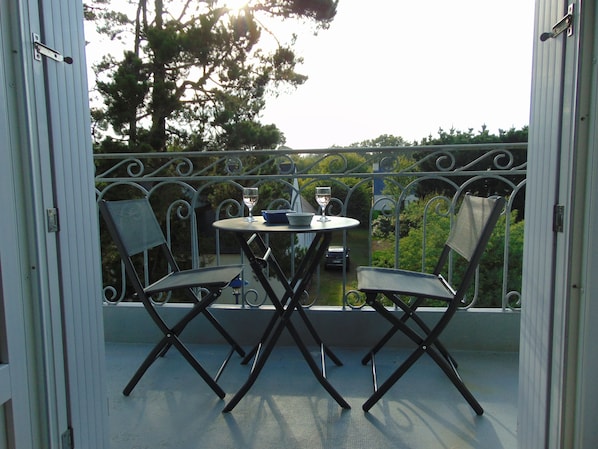 Een balkon met een tafel en twee stoelen, een auto op de achtergrond, en uitzicht op bomen en een gebouw.