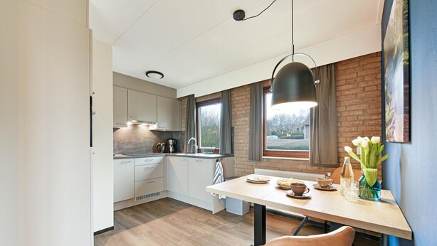 A modern kitchen with white cabinets, a brick accent wall, and a dining area with a table and chairs. - South Limburg Stay Near Tripoint (Vijlen)