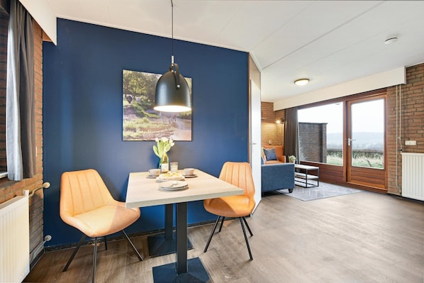 Un coin repas avec une table et deux chaises orange, un luminaire suspendu et une grande fenêtre donnant sur un beau paysage.