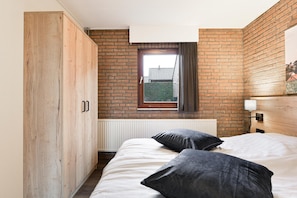 Une chambre avec un mur en briques, une fenêtre avec des rideaux, une armoire en bois et un lit avec des oreillers.