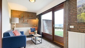 A modern living room with a blue sofa, a small coffee table, and a view of a green landscape through a large window. - South Limburg Stay Near Tripoint (Vijlen)