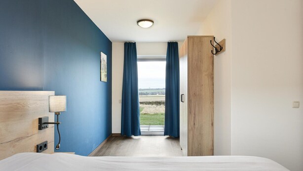 A bedroom with a bed, a nightstand, a lamp, a wooden wardrobe, and a view through a window with curtains. - Holiday Home Near Vijlen (Vijlen)