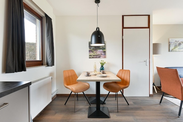 A dining area with a table, two chairs, a pendant light, a window with curtains, a radiator, and a door.