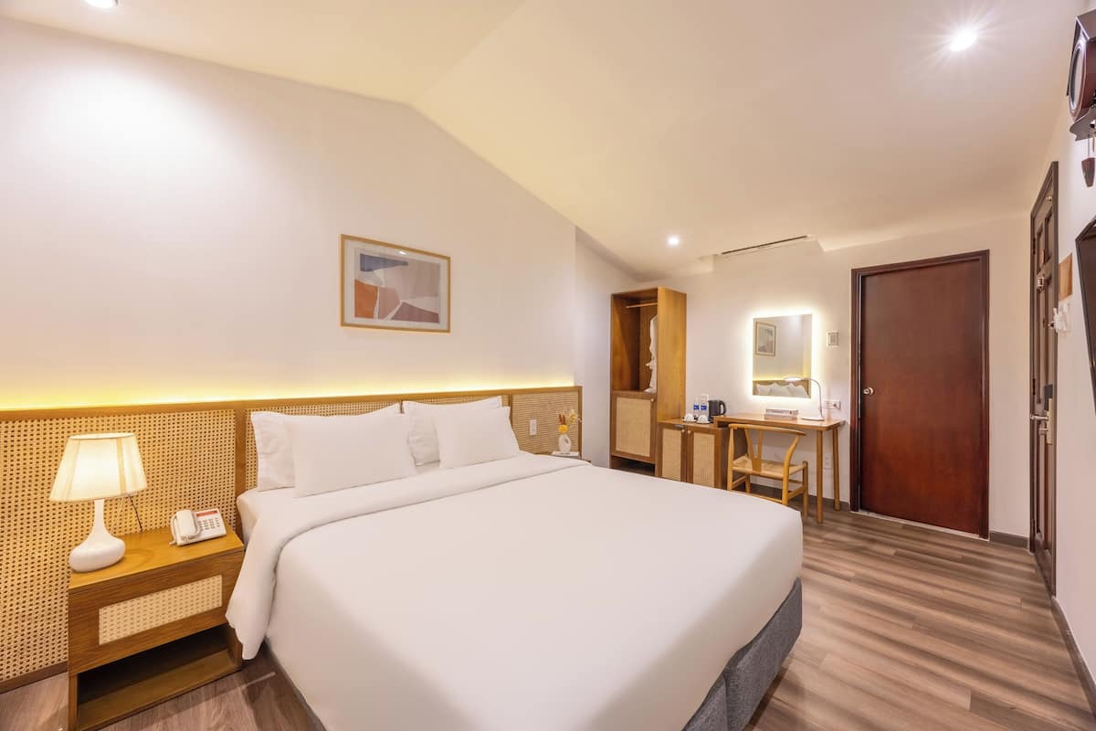 Ein Hotelzimmer mit einem großen Bett, einem Nachttisch mit Lampe, einem kleinen Schreibtisch mit Stuhl, einem Holzschrank und einer Tür.