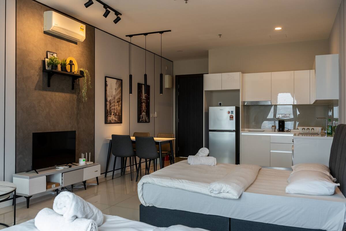 A modern hotel room with a TV, a small dining area, a kitchen, and a bed with towels.