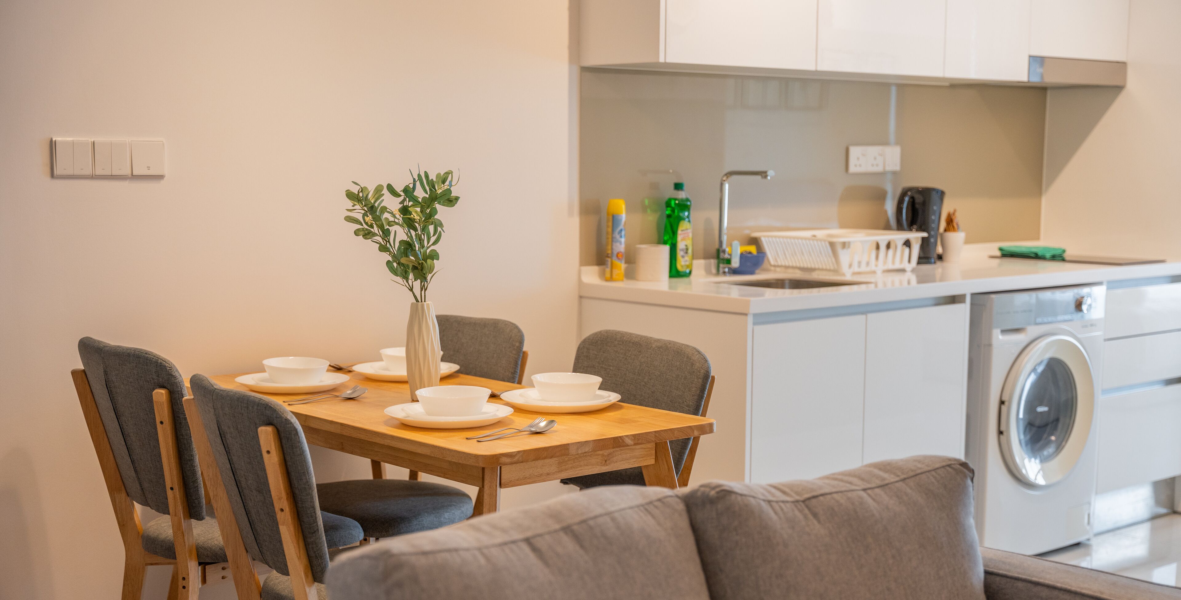 Una cocina moderna con zona de comedor, que incluye una mesa de madera preparada para cuatro personas, sillas tapizadas en gris y un sofá de color gris. Se observa una lavadora al fondo.