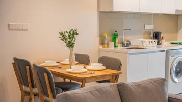 Una cocina moderna con zona de comedor, que incluye una mesa de madera preparada para cuatro personas, sillas tapizadas en gris y un sofá de color gris. Se observa una lavadora al fondo.