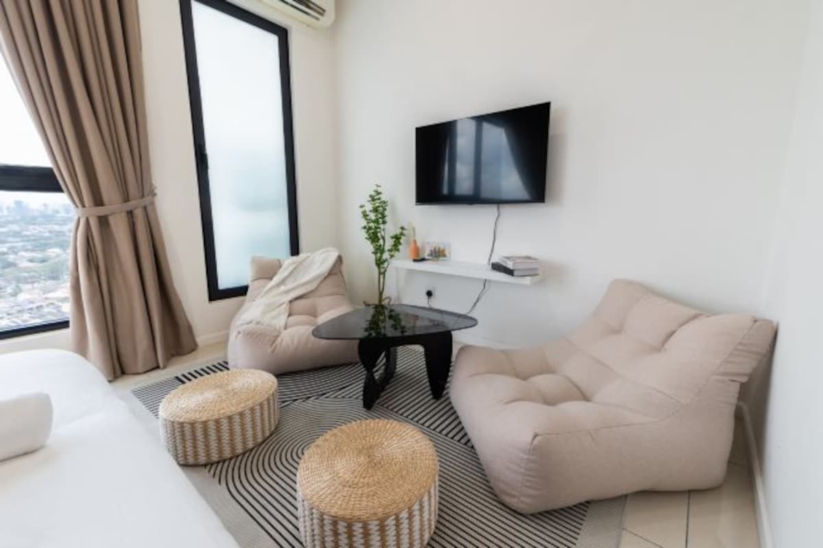 A modern hotel room with a large window, a flat-screen TV, a sofa, a coffee table, and two ottomans.