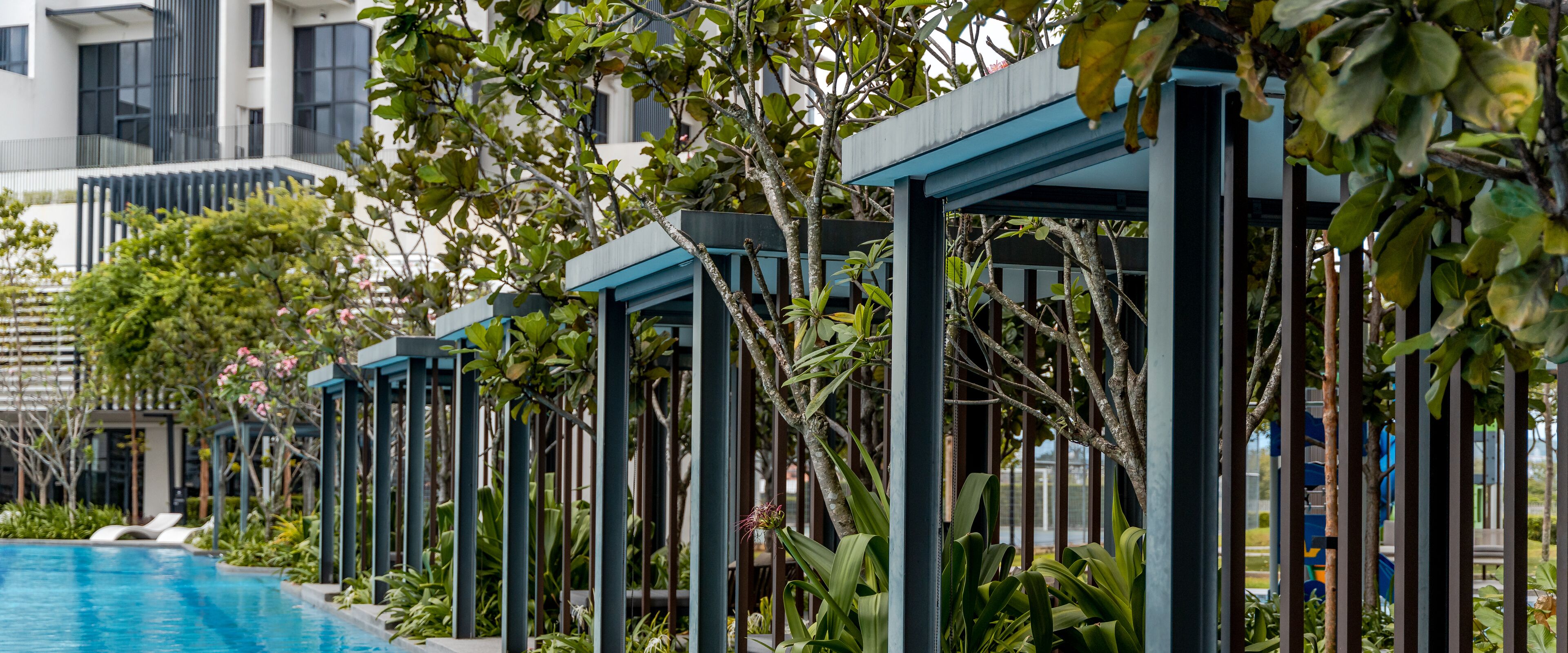 A poolside area with a series of covered seating areas, surrounded by lush greenery and a modern building in the background.