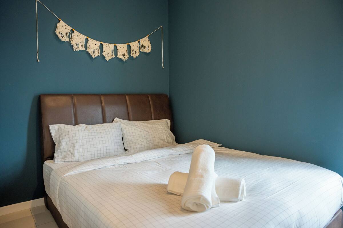 A neatly made bed with a rolled white towel, a brown headboard, and a macrame wall hanging.