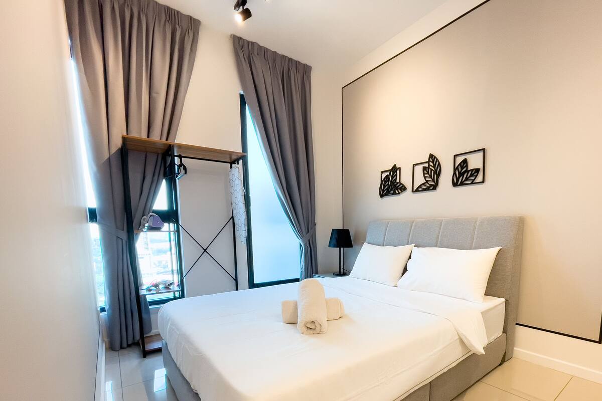 A neatly made bed with white linens and two rolled towels, a grey upholstered headboard, and a wall with three leaf-patterned wall art pieces.