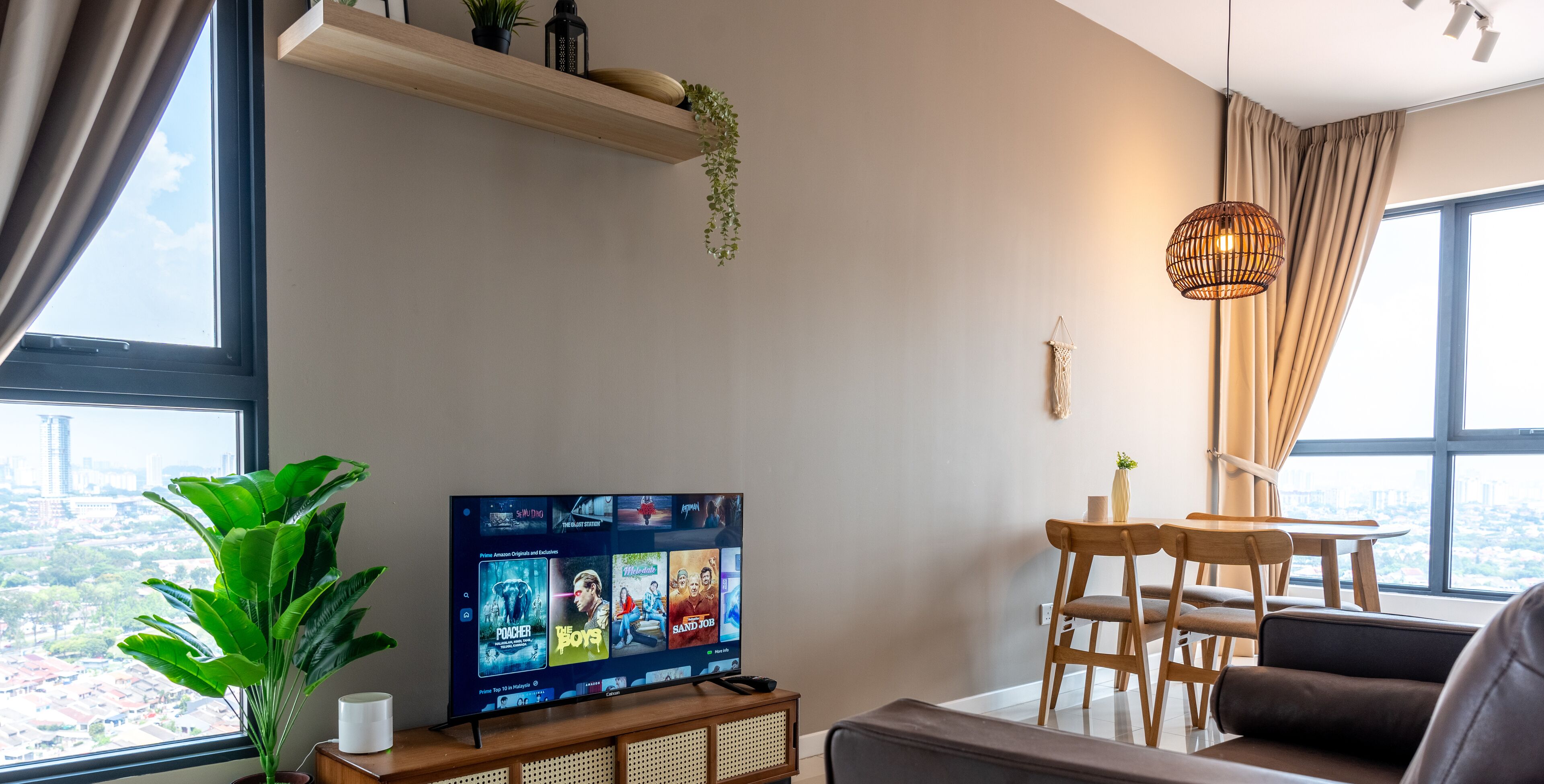 A modern living room with a TV displaying movie options, a potted plant, a wooden table, and a view of the city through large windows.
