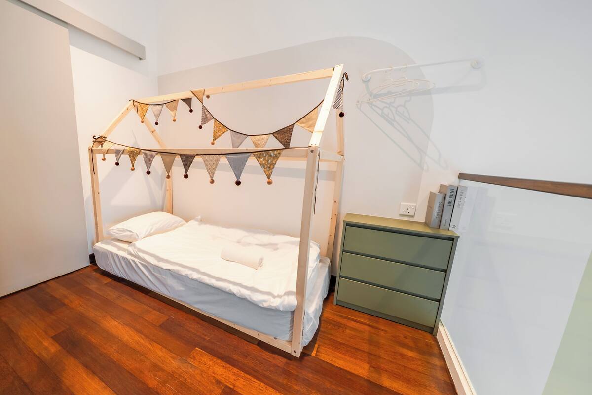 A single bed with a wooden frame and a canopy, a green nightstand, and a white wall with a power outlet.