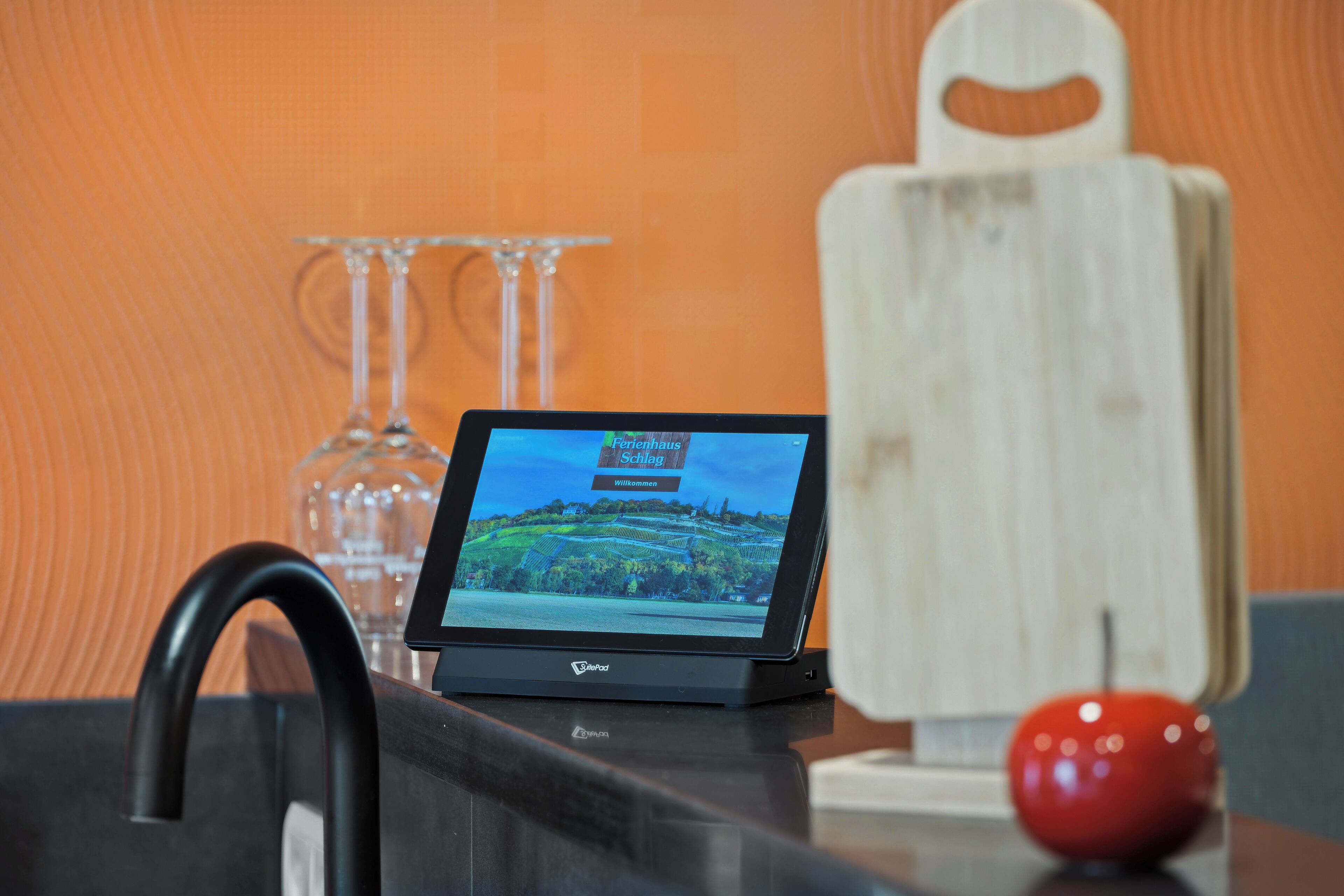 Een tablet toont een welkomstscherm met de tekst ‘Ferrenhaus Willkommen’ op een aanrecht met een zwarte kraan, een rode appel en een houten snijplank.