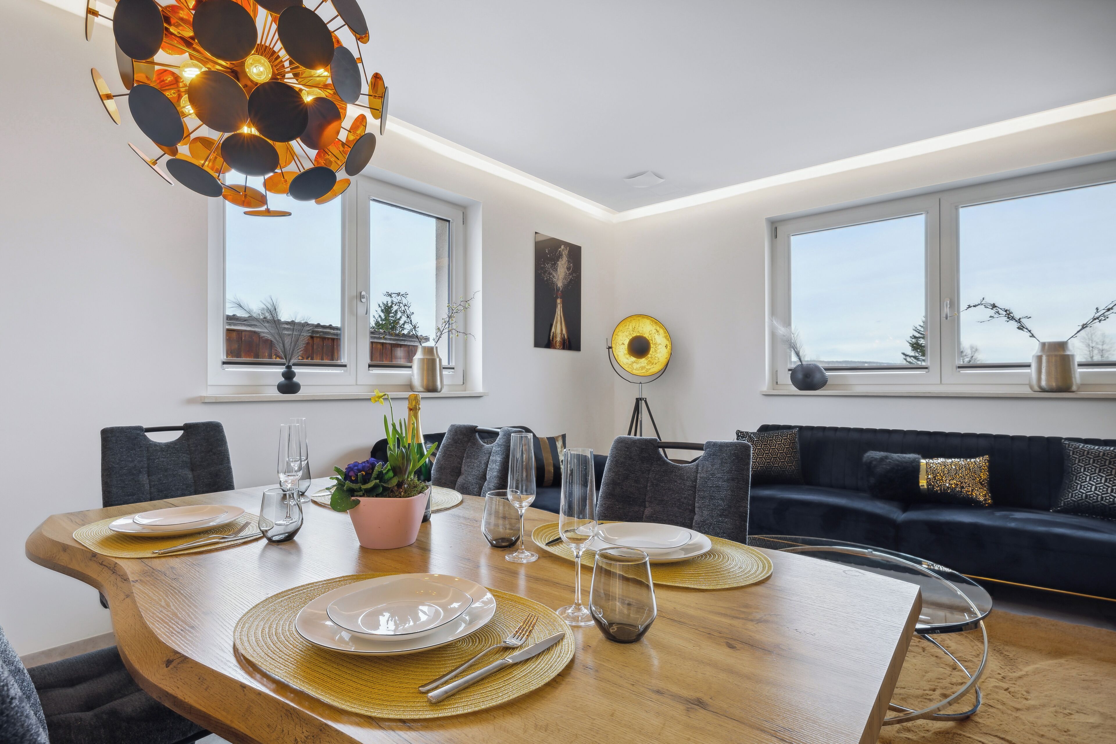 A dining area with a wooden table set for four, surrounded by grey chairs, a dark sofa, and a modern pendant light.